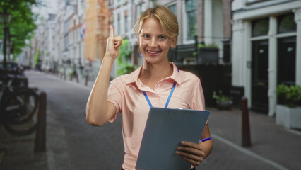 Woman smiling while holding a blue clipboard and pointing finger toward it on a street lined with...