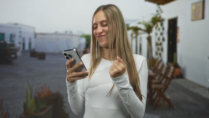 Young woman texting on smartphone with clenched fist on a sunlit street near outdoor chairs and potted plants; joy success.