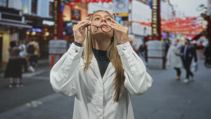 Woman adjusting glasses with hands, face visible, standing in a busy city street near shops and lanterns; quiet contemplation.