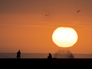 Naklejka premium Silhouetted people at a coastal breakwater during a dramatic sunset with crashing waves and birds in flight