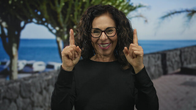 Woman pointing index fingers upward on seaside promenade by ocean; optimism hope positivity inspiration.