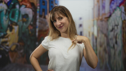Fototapeta premium Woman points finger to chest while smiling on graffiti covered street in a white t shirt and relaxed pose; confidence.