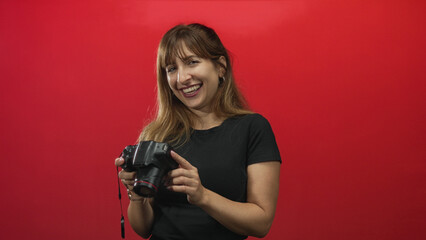 Woman smiling while holding a dslr camera and checking images in a studio against a red wall; creative joy.
