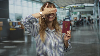 Woman holding passport with hand covering eyes at a busy airport terminal, smiling broadly while...