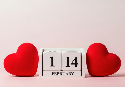 Two plush red hearts flank a wooden calendar displaying february 14th on a soft pink background, representing day