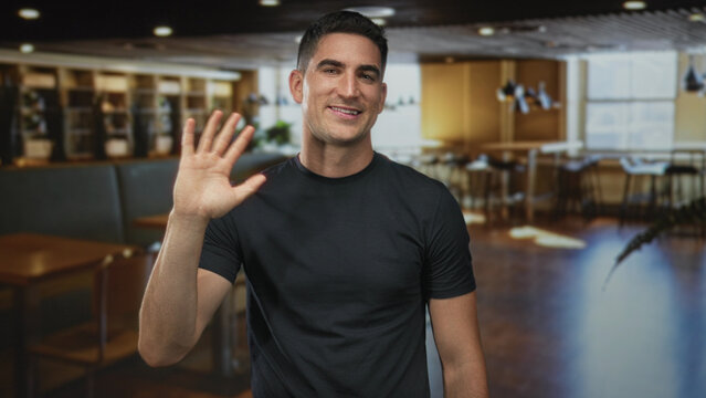 Man waving hand in building lobby with bare forearm visible and wearing black t shirt while smiling to camera; friendly greeting.