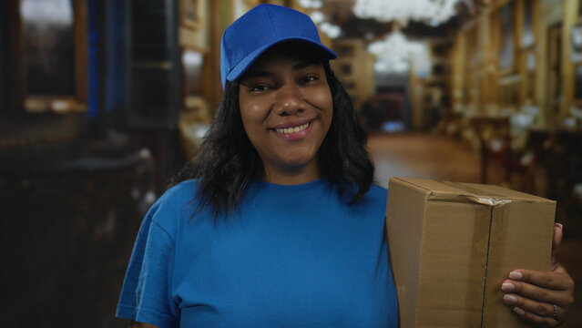 Smiling woman wearing cap in blue uniform holds large cardboard box with hand in building; friendly service.