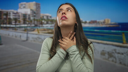 Fototapeta premium Young hispanic woman at seaside promenade holding throat in distress, surrounded by ocean and urban backdrop, underscoring tension in an outdoor setting.