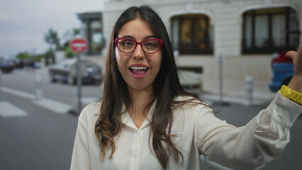 Young hispanic woman sends a kiss gesture on a bustling street outside a building; playful...
