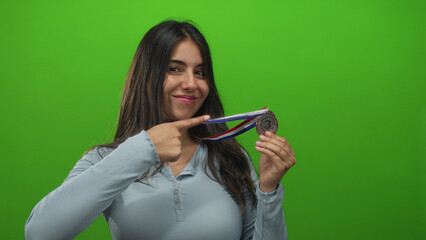 Young hispanic woman smiles while holding a bronze medal with thumb raised against a green studio wall; confidence success achievement pride.