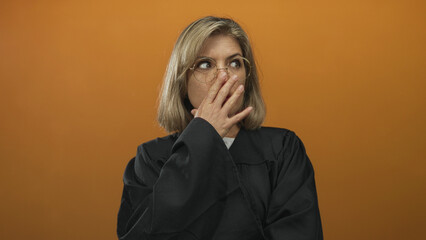 Young blonde judge wearing black robe covering mouth with finger to lips in orange studio; quiet...