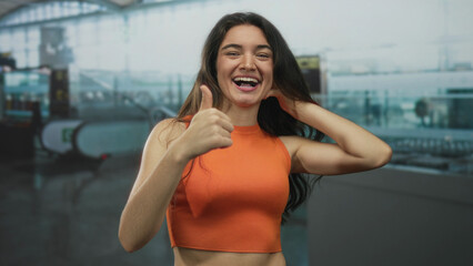 Young hispanic woman gives thumbs up gesture at airport terminal in orange top; optimism...