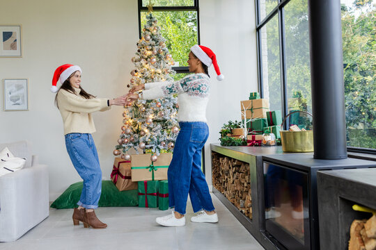 Diverse mother and daughter wearing Santa hats spinning by Christmas tree in living room with gifts