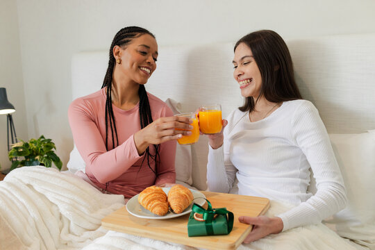 Diverse female couple sitting and clinking juice glasses over gift box on tray on bed - Powered by Adobe