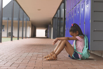 Child girl sitting on brick walkway leaning against purple lockers with teal backpack, copy space