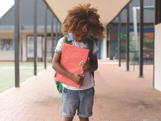 Female child standing, holding red binder to chest, looking down on covered brick walkway at school