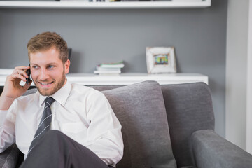 Adult man sitting on sofa in living room holding smartphone and wearing striped tie, copy space