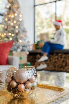 Bowl holding baubles on tray, tree shining with lights beside gifts, Santa hat wearer using device