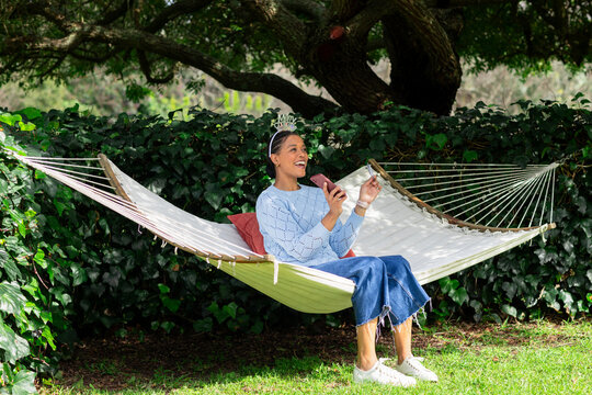 African American woman sitting on hammock in garden with tiara holding sparkler checking smartphone
