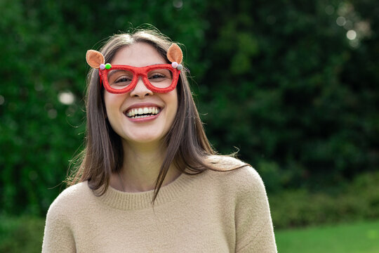 Woman smiling while standing outdoors in garden wearing beige knit sweater and red novelty glasses