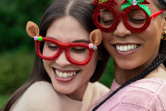 Female friends standing hugging each other in park wearing reindeer glasses and knit sweaters
