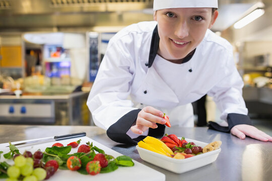 Female chef wearing white chef coat leaning over counter in kitchen placing strawberry into dish