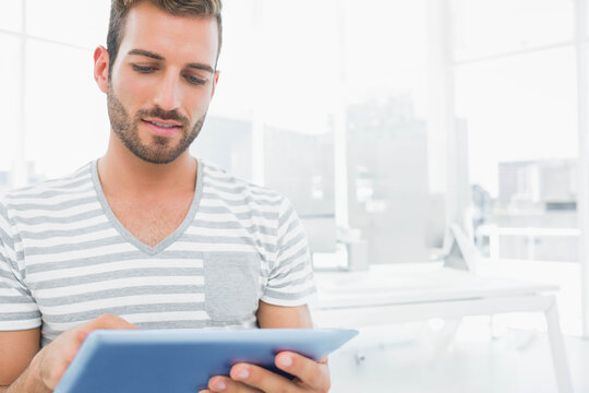 Bearded man wearing striped tee using light-blue tablet by white desks and monitors in office - Powered by Adobe