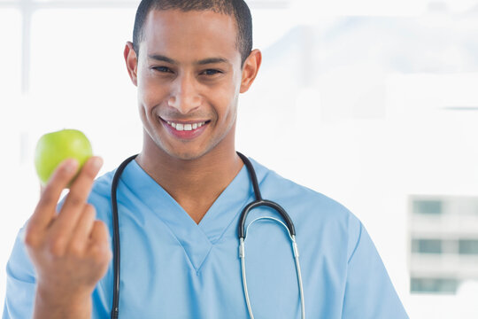 African American male in light blue scrubs holding green apple smiling in clinic with stethoscope - Powered by Adobe