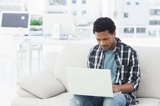 African American man typing on white laptop on beige sofa in bright workspace, copy space