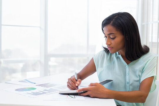 African American woman writing on notebook with charts at desk wearing mint blouse, copy space