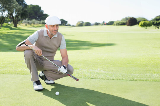 Mature man crouching, lining up putt on putting green with putter, ball, glove, cap, copy space