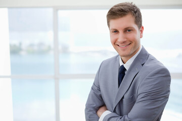 Man standing arms crossed at office windows overlooking waterfront in gray suit and tie, copy space