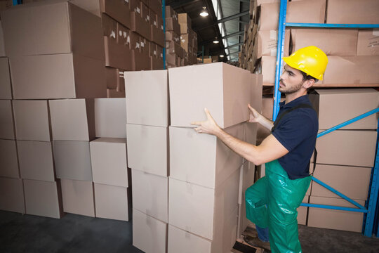 Male warehouse worker in yellow hard hat placing cardboard box onto stack near racking, copy space
