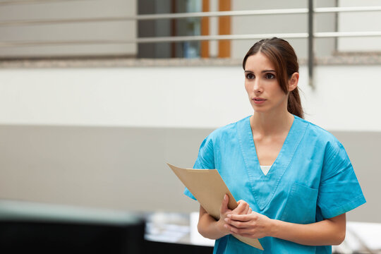 Woman in teal scrubs standing in clinical corridor holding manila folder by low counter, copy space