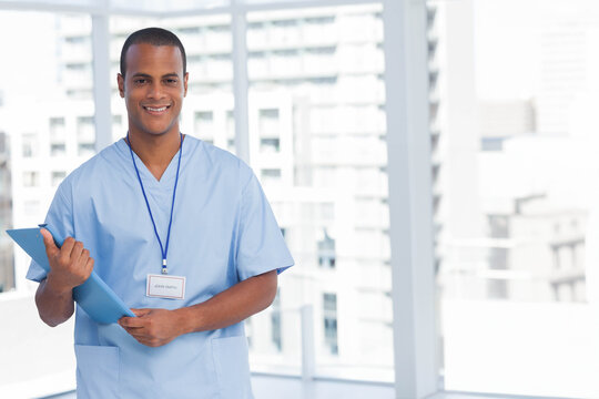 African American male nurse standing holding blue clipboard in lobby wearing blue scrubs ID badge - Powered by Adobe