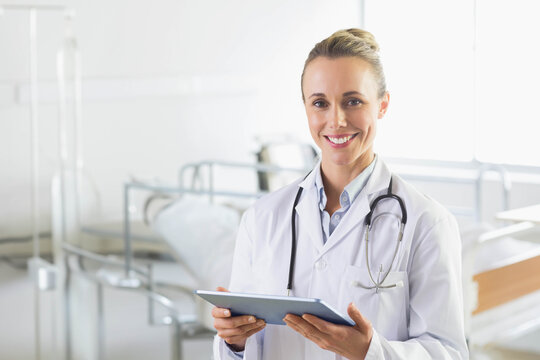 Midlife woman doctor wearing lab coat standing in ward holding tablet and stethoscope, copy space