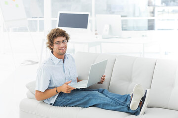 Man reclining on white leather sofa in bright lounge using silver laptop, wearing light-blue shirt