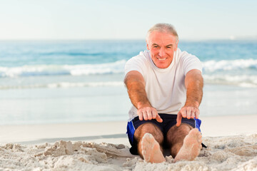 Naklejka premium Senior man sitting and stretching on sandy beach near ocean waves wearing short-sleeve T-shirt