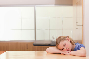 School-age girl resting on folded arms at kitchen table, looking at camera, blue top, copy space