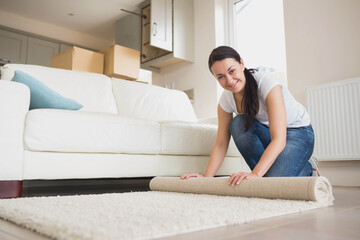 Adult female kneeling in living room rolling beige rug near sofa with moving boxes, copy space