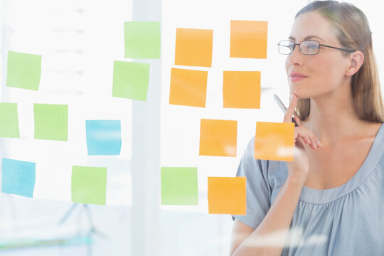 Middle-aged woman studying sticky notes on glass at office in blouse holding pen, copy space