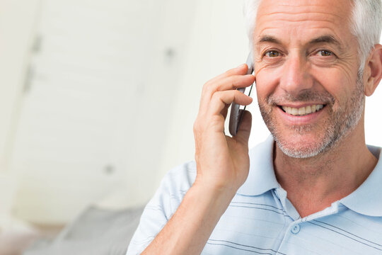 Senior man holding smartphone, smiling and speaking in bright room with light blue polo, copy space
