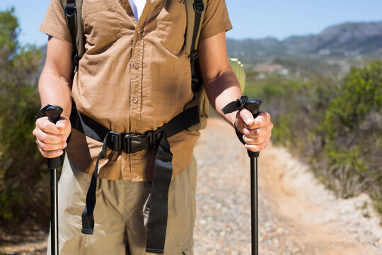 Male hiker standing on trail holding black trekking poles, wearing brown shirt and backpack - Powered by Adobe