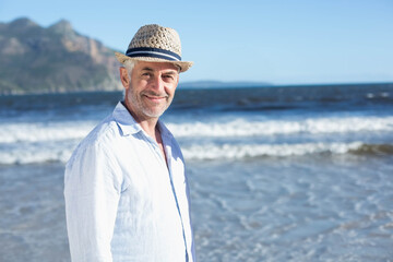 Man standing in shallow water at sandy beach wearing linen shirt and straw hat, looking