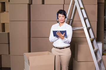 Mature woman in white shirt holding tablet wearing headset, checking open box in warehouse stacks