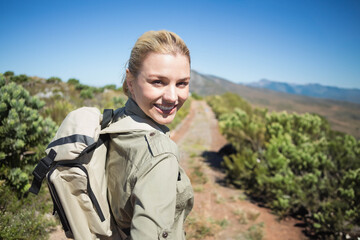Female hiker walking narrow trail in coastal scrub, wearing khaki shirt, carrying beige backpack