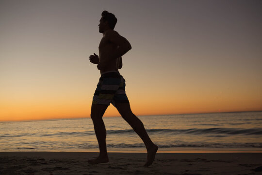 Shirtless man jogging along beach at sunrise wearing striped swim shorts leaving footprints - Powered by Adobe