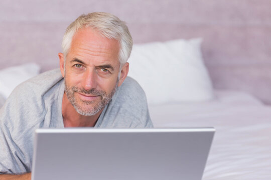 Mature man propping elbows, lying on bed using silver laptop in bedroom wearing grey T-shirt