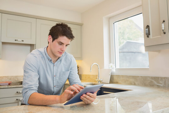 Man sitting at kitchen counter leaning forward using tablet near sink and red notebook, copy space - Powered by Adobe