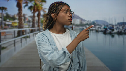 Woman points finger to boats on a street pier beside a marina walkway while wearing glasses and...
